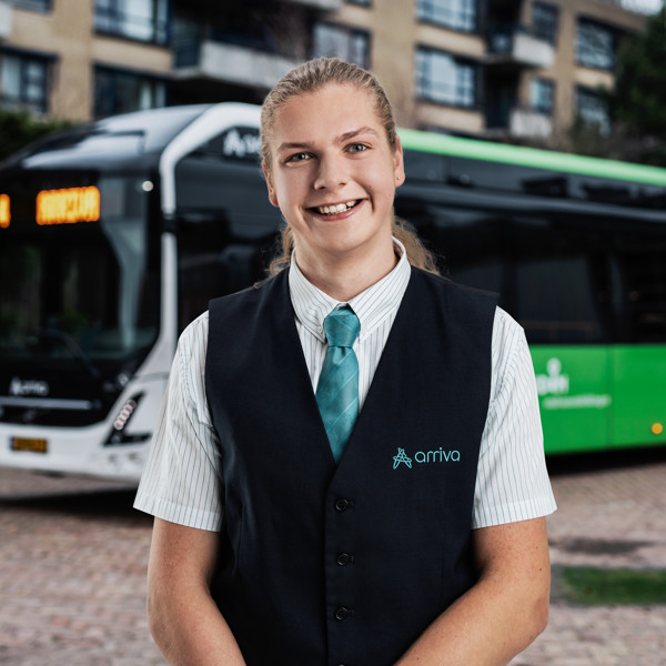 Buschauffeur Jarno heeft zijn haar in een staart naar achter en staat in Arriva-uniform lachend voor zijn bus.
