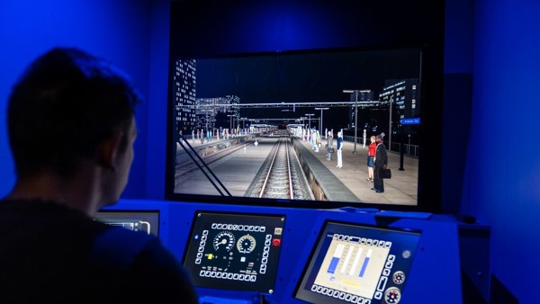 Een man zit in een treinsimulator, hij kijkt naar een groot scherm met daarop het spoor en heeft zijn handen op de stuurhendels liggen.
