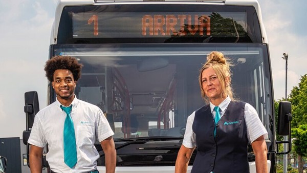 Buschauffeurs Griffin en Vera staan in hun Arriva-uniform lachend voor hun bus waarop staat '1 Arriva'.