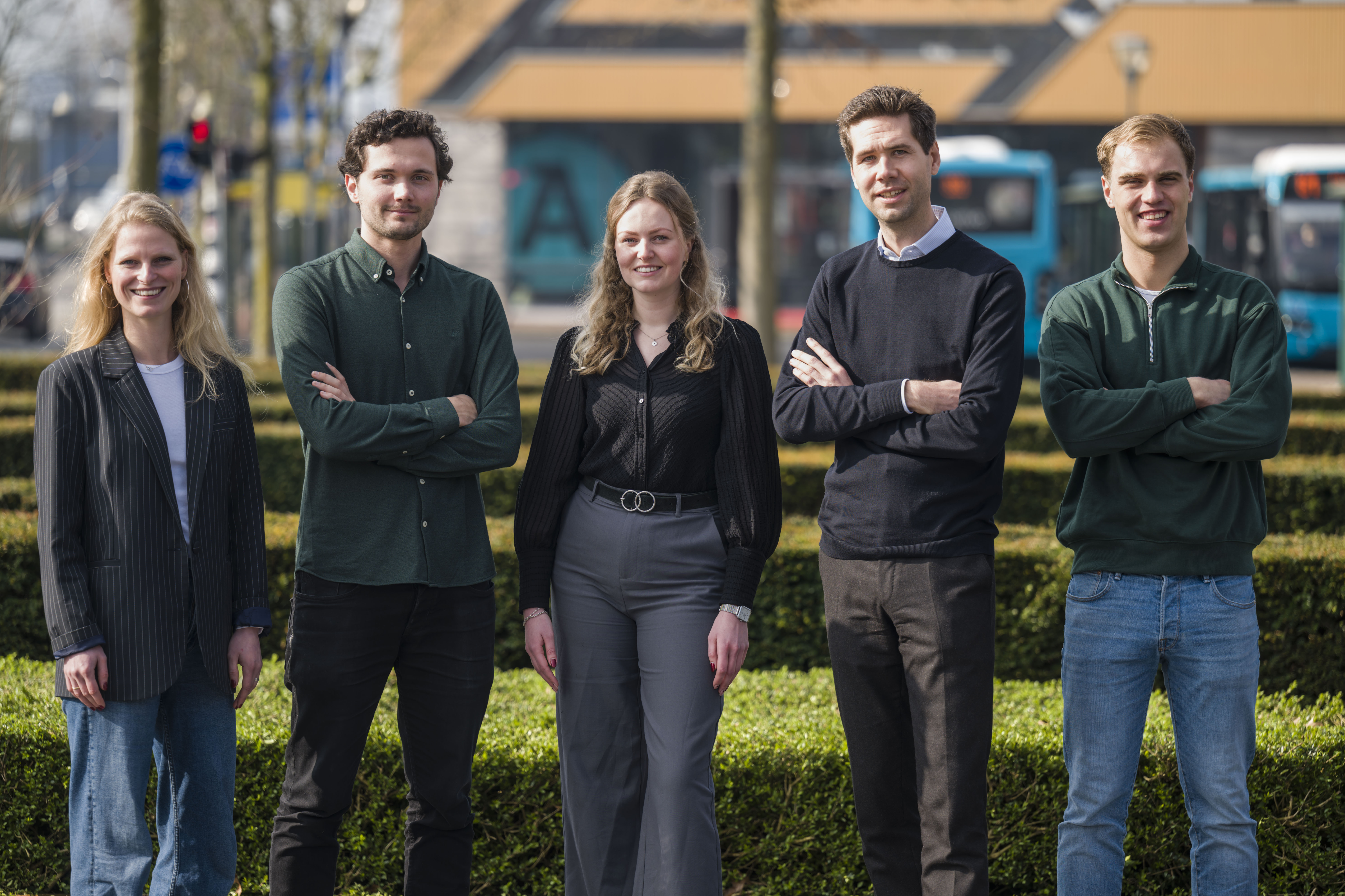 Vijf trainees, 2 vrouwen en 3 mannen, staan zelfverzekerd buiten tegen en stedelijk decor met bomen en Arriva-bussen. 
