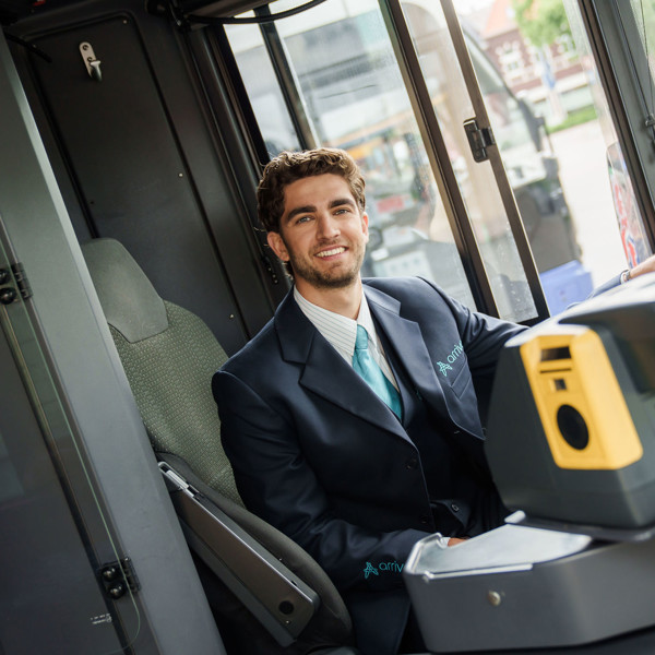 Een lachende buschauffeur in Arriva-uniform zit achter het stuur van een bus en kijkt naar de camera. Een lachende buschauffeur in Arriva-uniform zit achter het stuur van een bus en kijkt naar de camera.