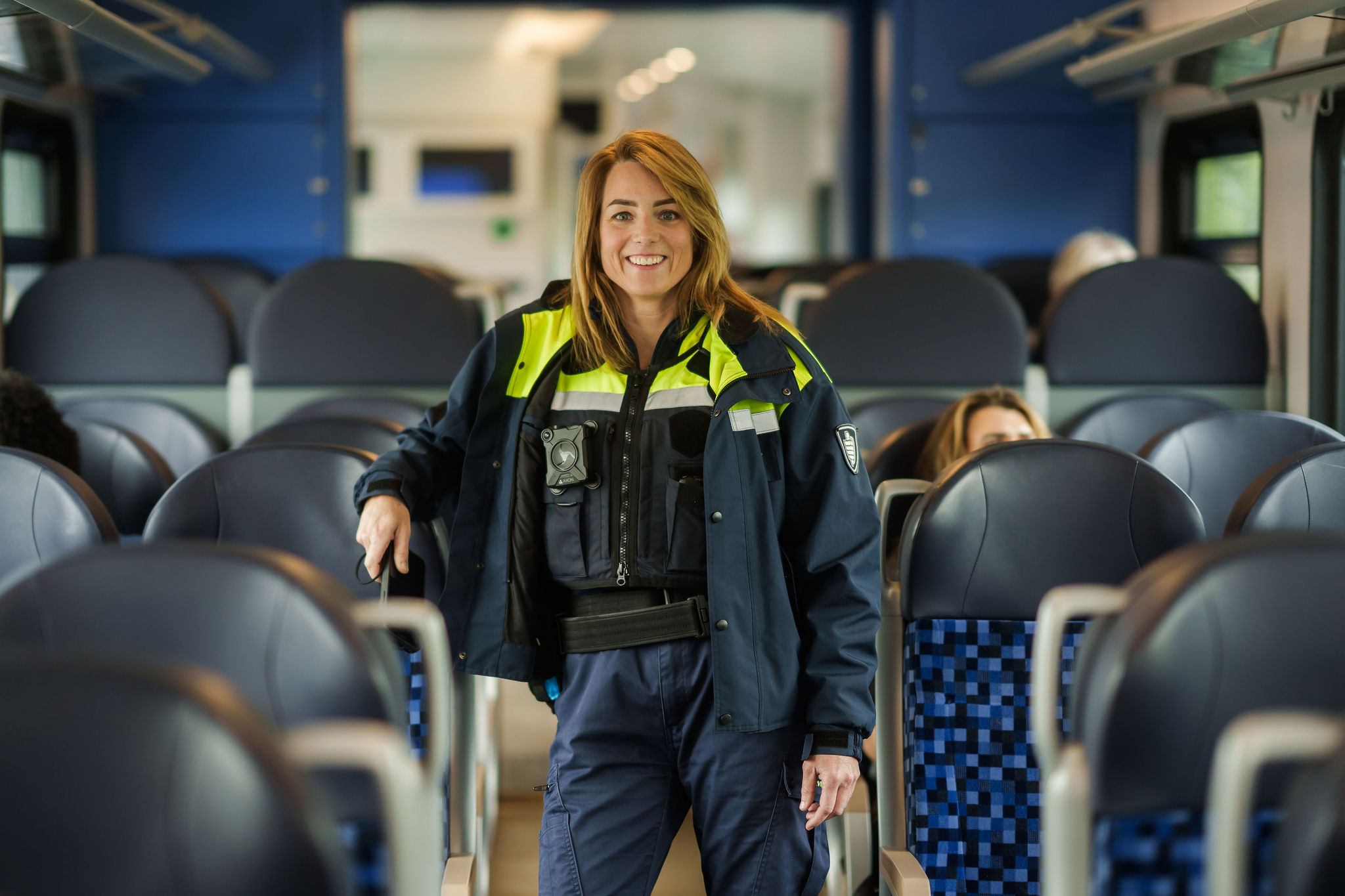 Een glimlachende veiligheidsmedewerker in uniform staat in een trein tussen de stoelen. Er zijn een paar reizigers.