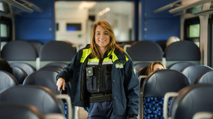Een glimlachende veiligheidsmedewerker in uniform staat in een trein tussen de stoelen. Er zijn een paar reizigers. Een glimlachende veiligheidsmedewerker in uniform staat in een trein tussen de stoelen. Er zijn een paar reizigers.