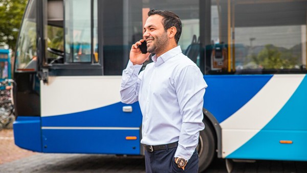 Teammanager Mohamed staat lachend te bellen buiten met een bus op de achtergrond.