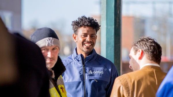 Een servicemedewerker van Arriva met zwart haar lacht naar de camera, om hem heen staan reizigers.