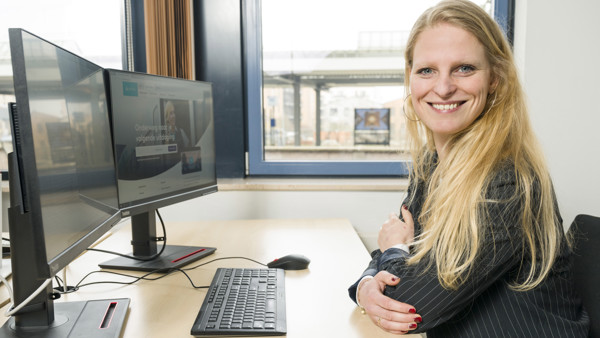 Vrouw in kantoor glimlacht naar camera, computerscherm met website Arriva zichtbaar, groot raam toont uitzicht op treinstation.