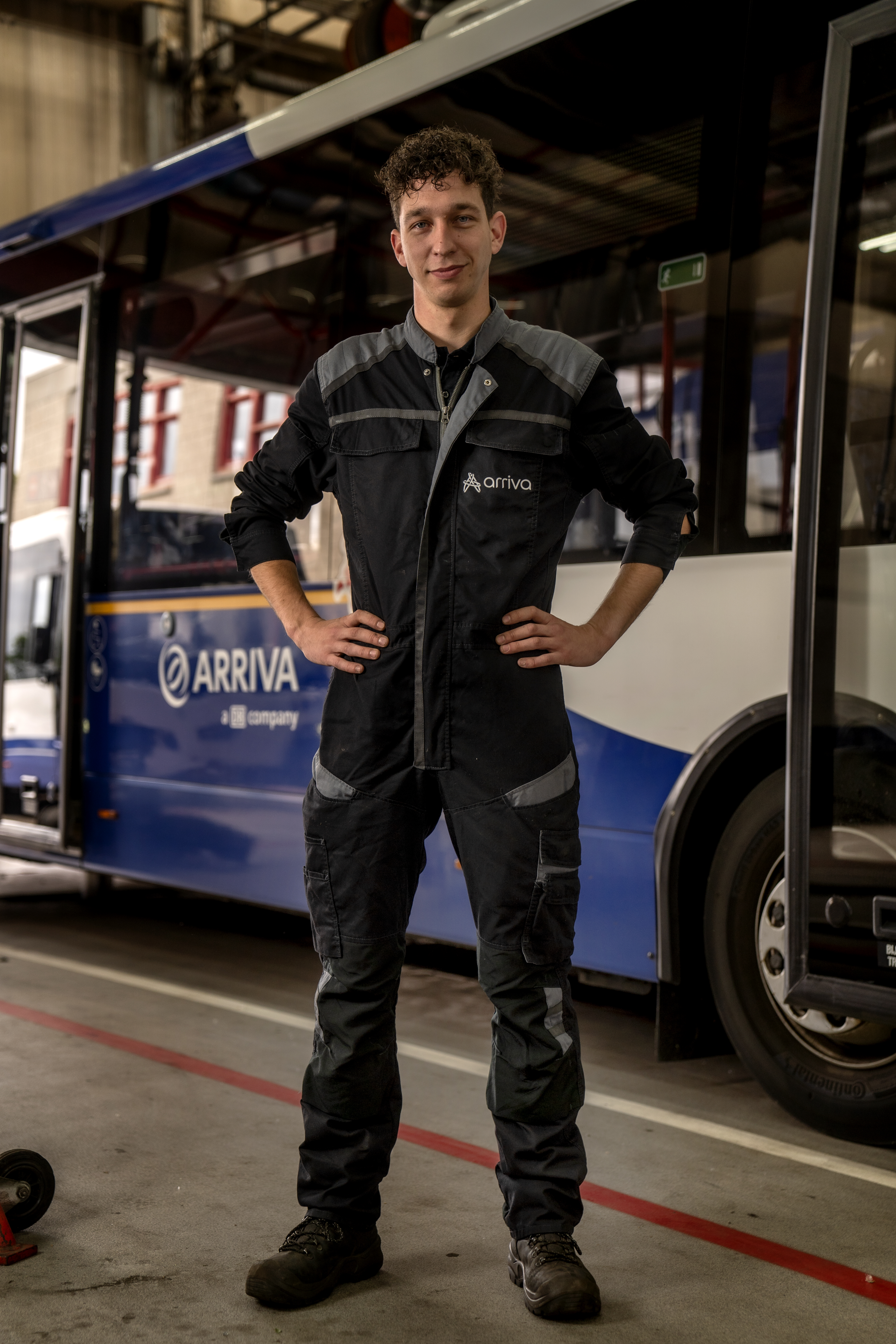 Mannelijke monteur, in werkplaatsuniform, kijkt stoer in de camera met handen in zijn zij. Een Arriva-bus op de achtergrond.