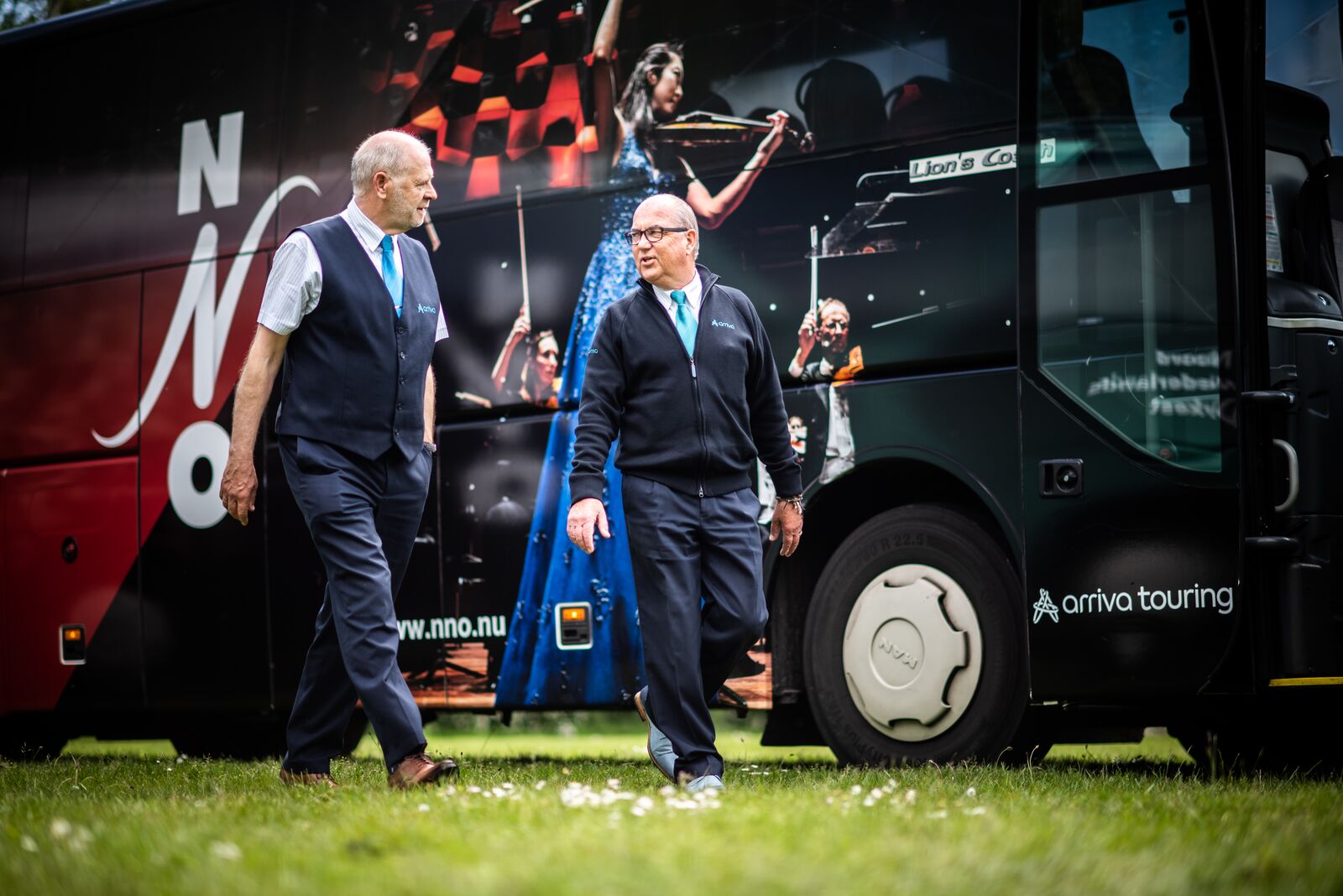  Twee mannelijke Arriva chauffeurs in uniform lopen langs een Arriva Touring bus, terwijl ze zich op een grasveld bevinden.