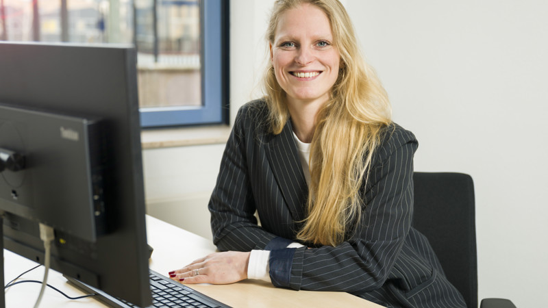Vrouw achter computer in kantoor, glimlacht naar camera in professionele kleding. Raam biedt uitzicht op trein en stadsgebouwen bij openbaar vervoer.