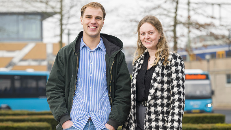 Man en vrouw buiten, glimlachend naar camera; hij in casual jas en gestreept shirt, zij in geruite jas, tegen stedelijke achtergrond met bus- en treinstation.