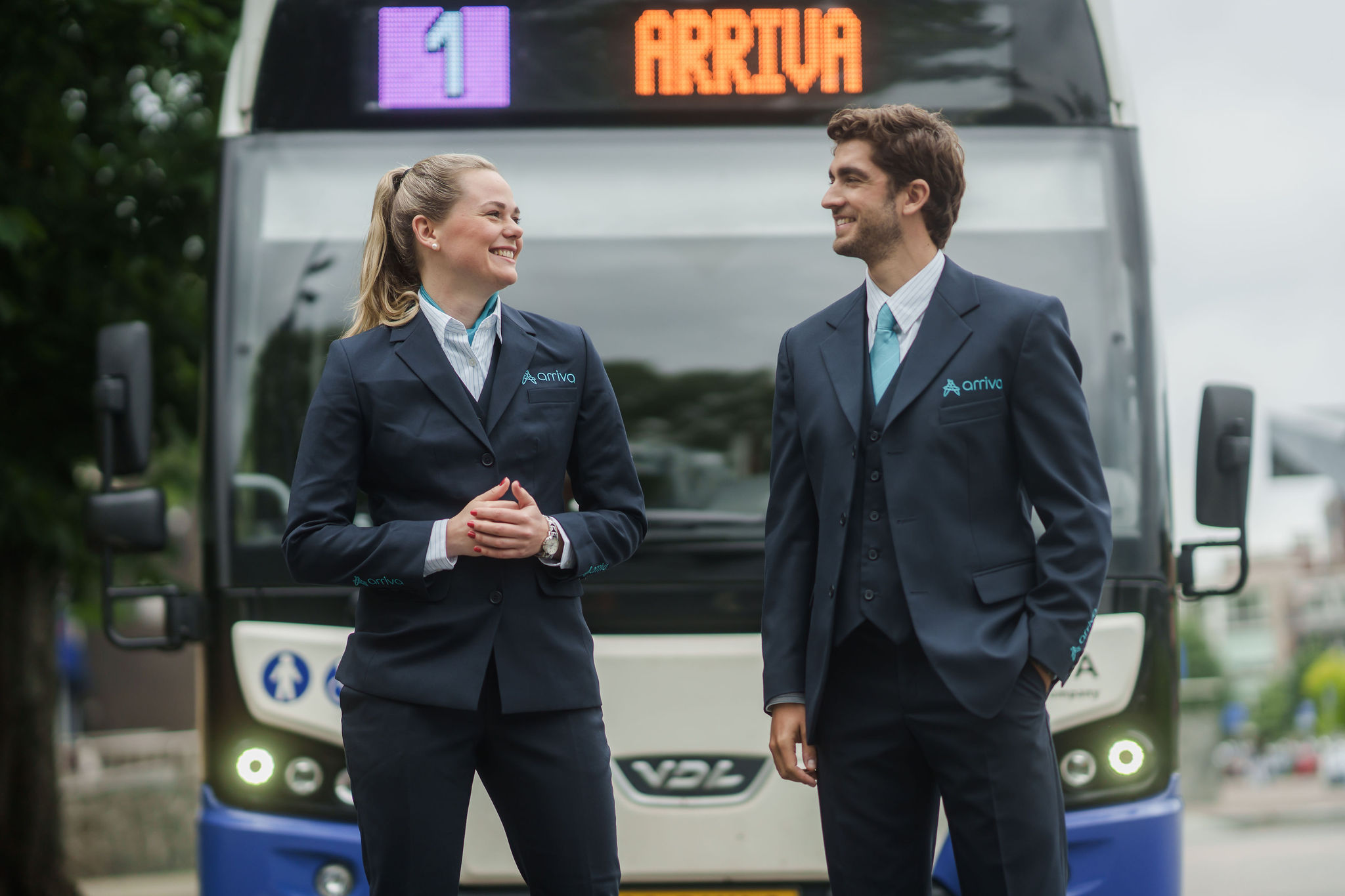 Twee lachende Arriva-buschauffeurs in uniform staan voor een bus met lijnnummer 1 en Arriva op de achtergrond.