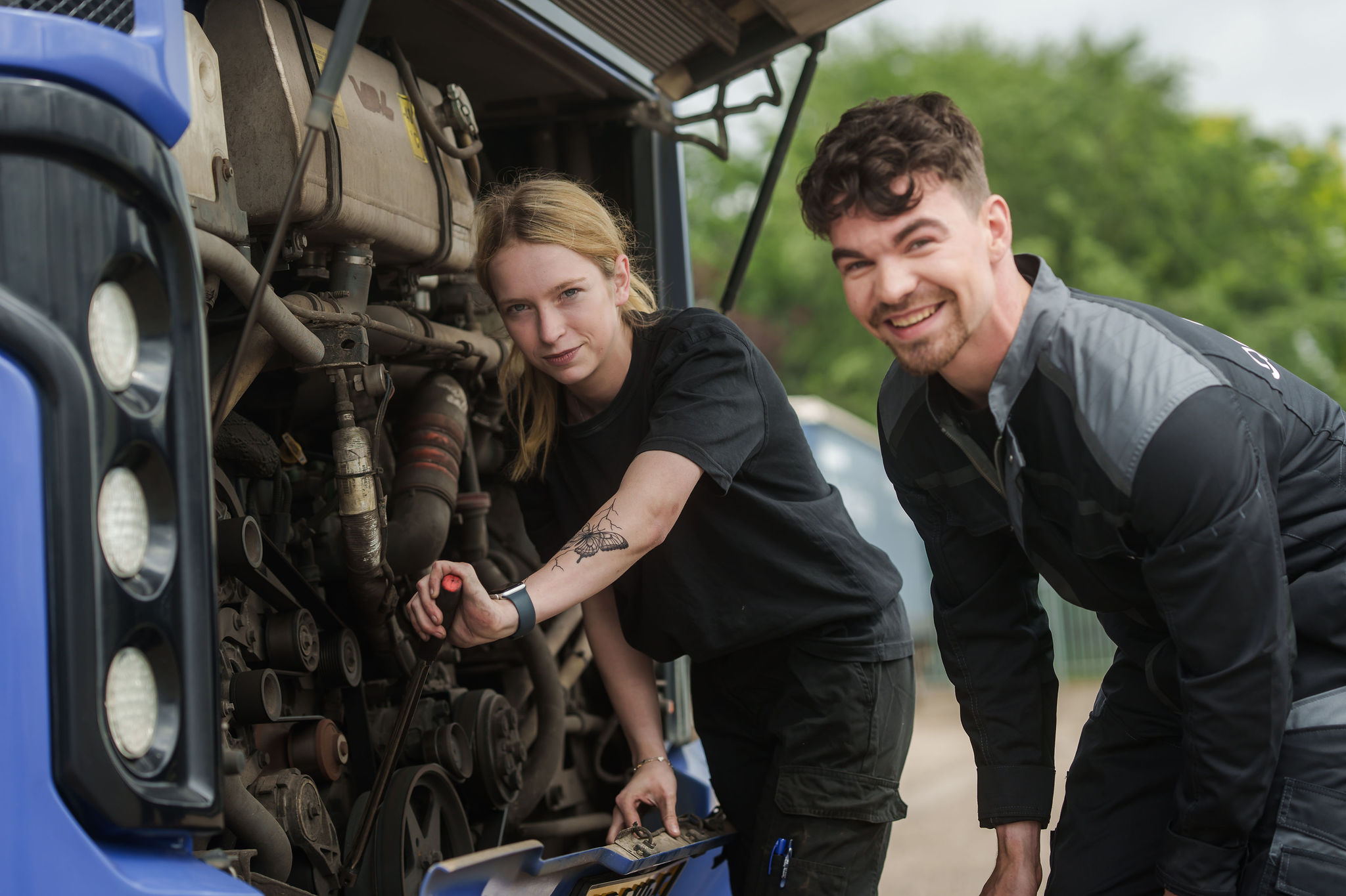 Twee monteurs werken aan een busmotor, zij sleutelt aan de motor en hij kijkt lachend in de camera. 
