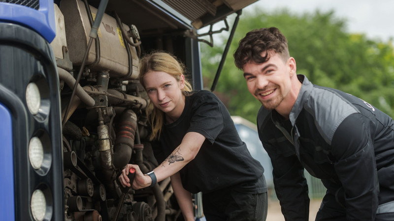 Twee monteurs werken aan een busmotor, zij sleutelt aan de motor en hij kijkt lachend in de camera. Twee monteurs werken aan een busmotor, zij sleutelt aan de motor en hij kijkt lachend in de camera.