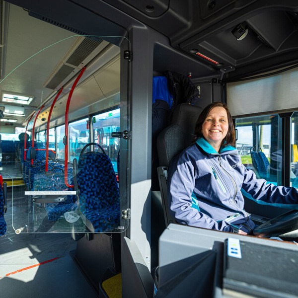 Een vrouwelijke buschauffeur met bruin haar zit achter het stuur van een Arriva-bus en glimlacht.