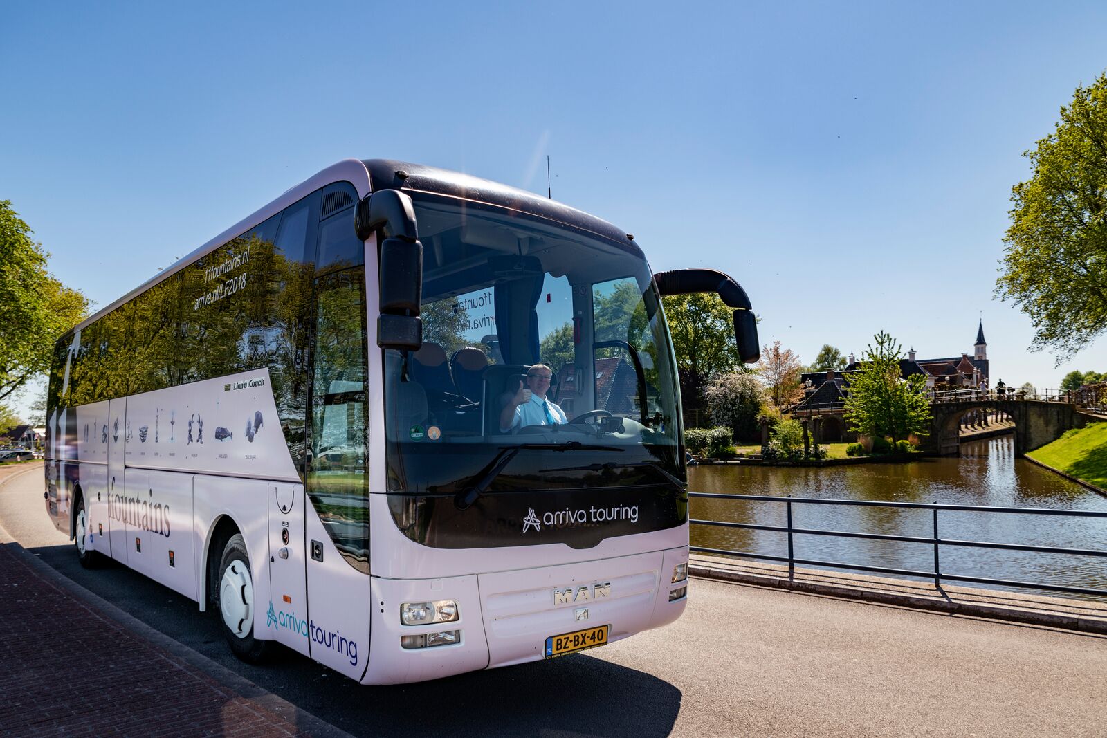Chauffeur rijdt grote witte Arriva-touringcar door een stadscentrum en steekt zijn duim op.