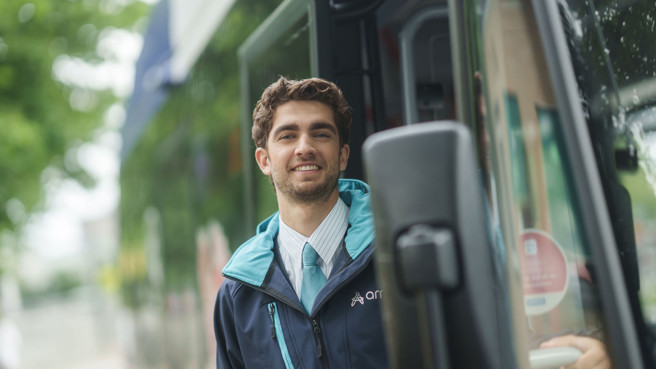 Een buschauffeur in Arriva-uniform met een jas staat glimlachend in de deuropening van een bus, met bomen op de achtergrond. Een buschauffeur in Arriva-uniform met een jas staat glimlachend in de deuropening van een bus, met bomen op de achtergrond.