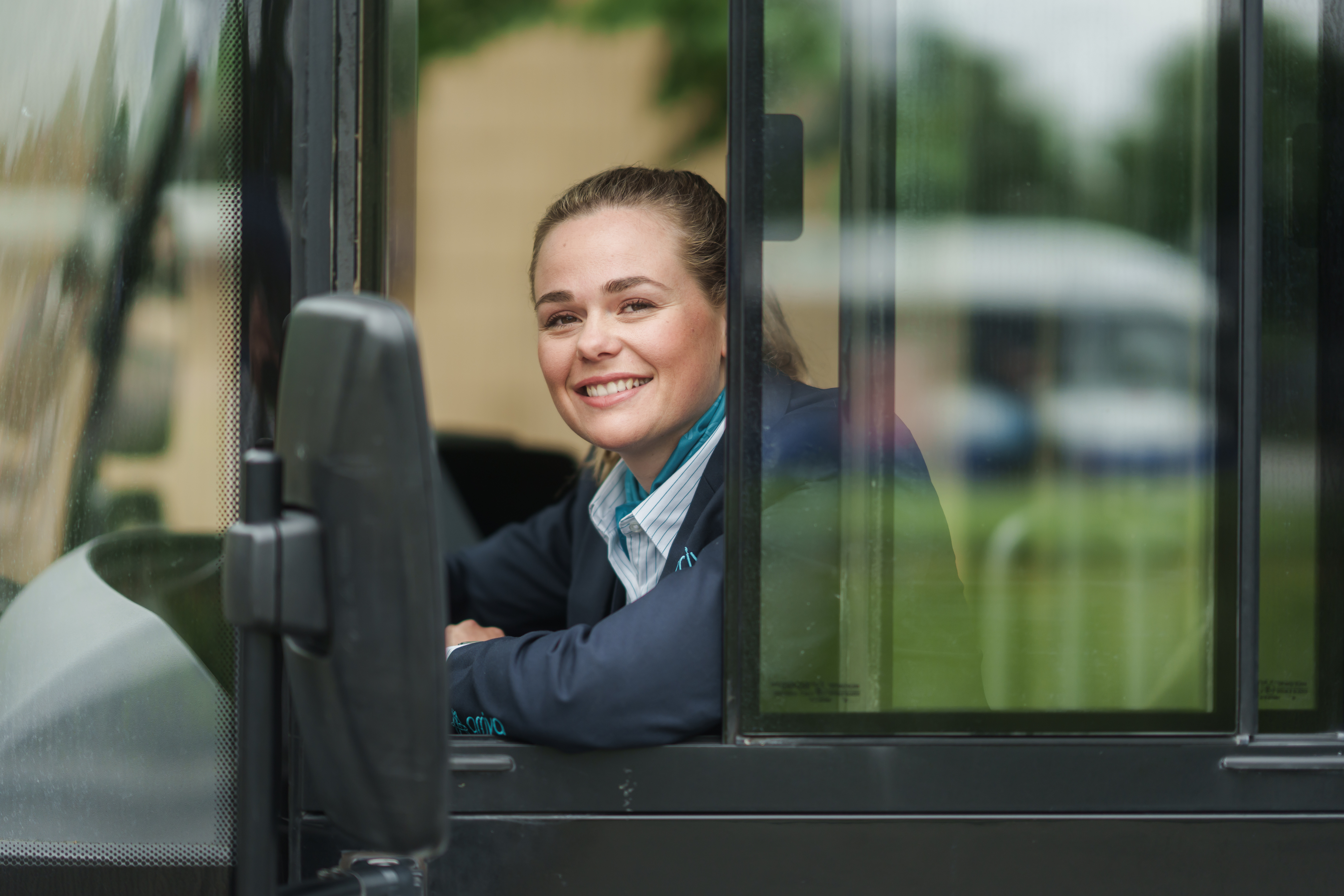 Arriva-chauffeur zit achter het stuur van haar bus en leunt relaxed uit haar zijraam en kijkt lachend in de camera.
