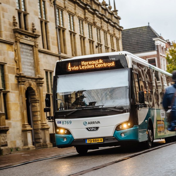Blauw-witte Arriva-bus rijdt door het centrum van Leiden langs een statig gebouw, met fietsers op de weg.
