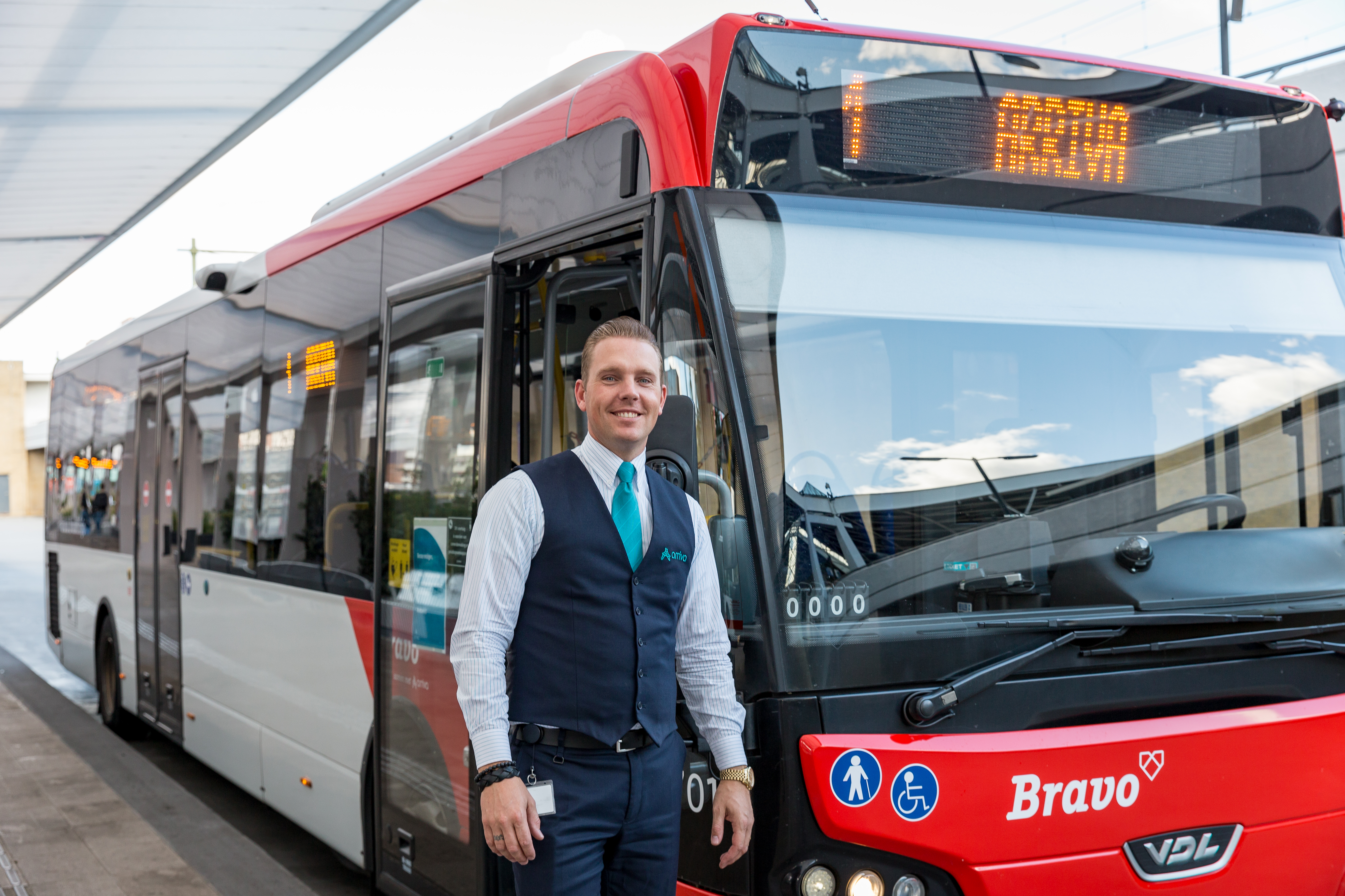 Een lachende chauffeur in uniform staat voor een rode bus van Arriva.