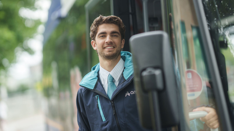 Een chauffeur van Arriva in uniform lacht naar de camera terwijl hij in de deuropening van zijn bus staat. Een chauffeur van Arriva in uniform lacht naar de camera terwijl hij in de deuropening van zijn bus staat.
