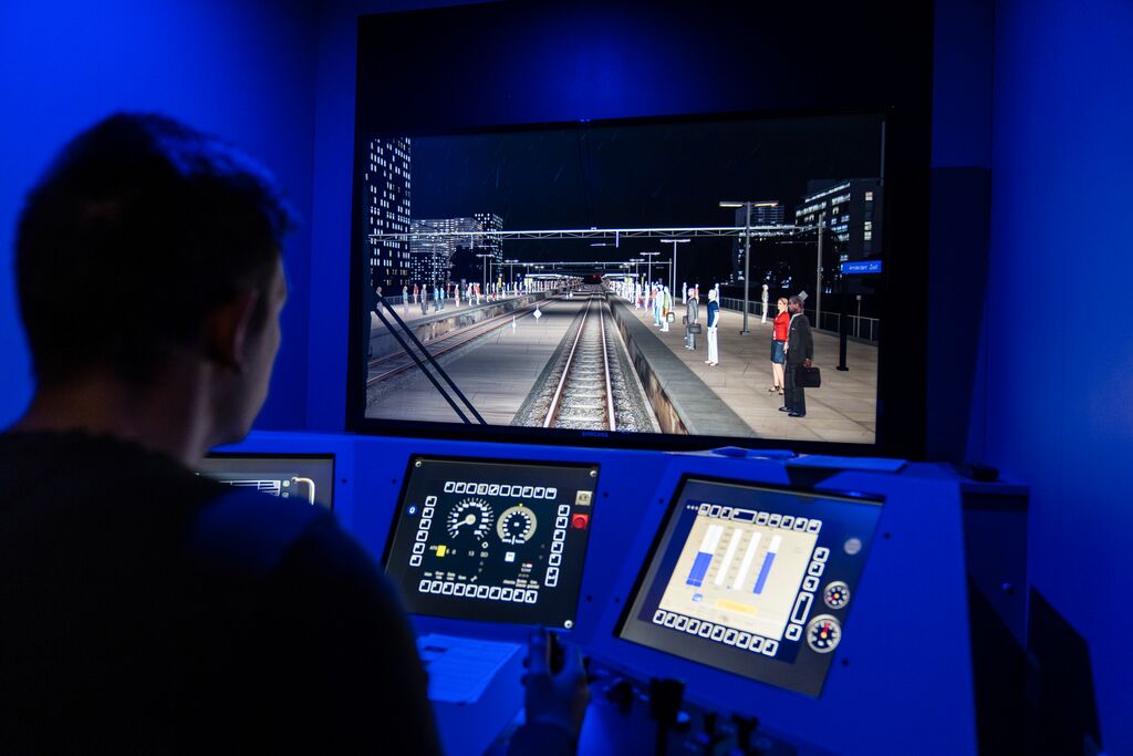Een man zit in een treinsimulator, hij kijkt naar een groot scherm met daarop het spoor en heeft zijn handen op de stuurhendels liggen.