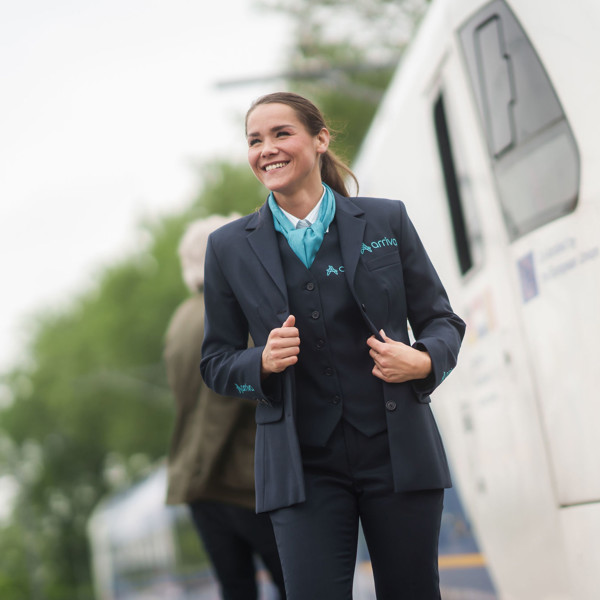 Een lachende machinist in Arriva-uniform staat buiten naast een trein, met bomen en een andere persoon op de achtergrond. Een lachende machinist in Arriva-uniform staat buiten naast een trein, met bomen en een andere persoon op de achtergrond.