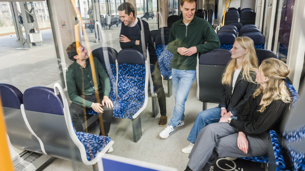 Vier trainees in gesprek in lege Arriva-trein, twee staan, twee zitten, ontspannen sfeer met natuurlijk licht door ramen.