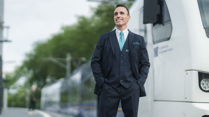 Machinist van Arriva glimlacht en kijkt in de verte met achter hem een trein. Machinist van Arriva glimlacht en kijkt in de verte met achter hem een trein.