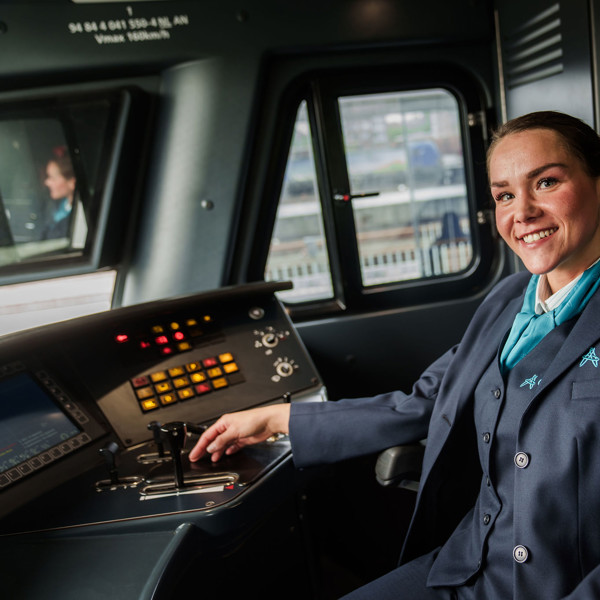 Een lachende machinist in Arriva-uniform zit in de bestuurderscabine van een trein en kijkt naar de camera. Een lachende machinist in Arriva-uniform zit in de bestuurderscabine van een trein en kijkt naar de camera.