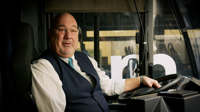 Buschauffeur Richard zit ontspannen voorin de bus. Zijn handen rusten losjes op het stuur en hij kijkt je terwijl hij tegen je praat.