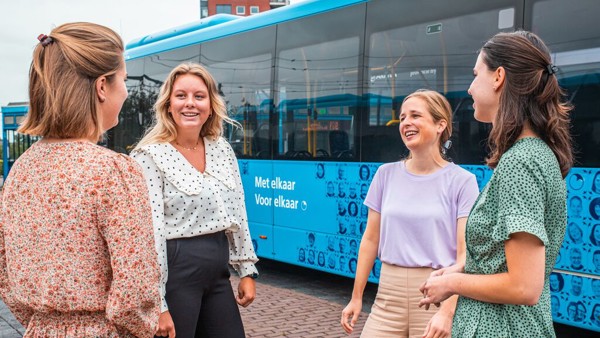 Vier jonge vrouwelijke collega's overleggen lachend met elkaar, op de achtergrond staan blauwe Arriva-bussen.