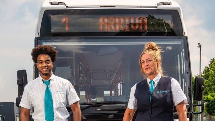 Buschauffeurs Griffin en Vera staan in hun Arriva-uniform lachend voor hun bus waarop staat '1 Arriva'.
