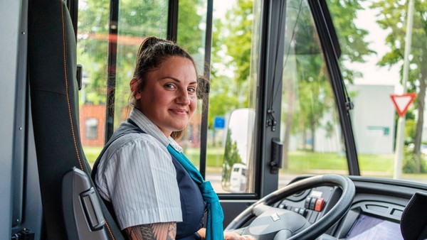 Buschauffeur Kelly van Arriva zit relaxed achter het stuur van haar bus en kijkt achterom naar haar reizigers.