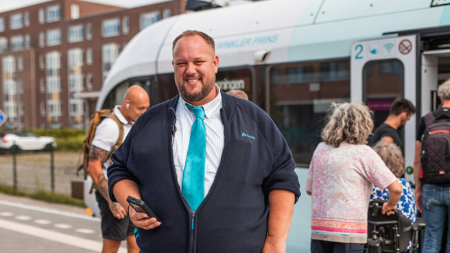 Steward Peter staat in zijn donkerblauwe Arriva vest en aquakleurige Arriva stropdas voor de trein. Achter hem stappen reizigers in. Hij gaat ze reisbewijzen controleren met het apparaat dat hij daarvoor in zijn werk gebruikt. 