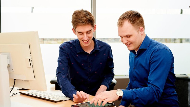 Twee jonge mannelijke collega's van Arriva zitten samen achter een computer op kantoor.