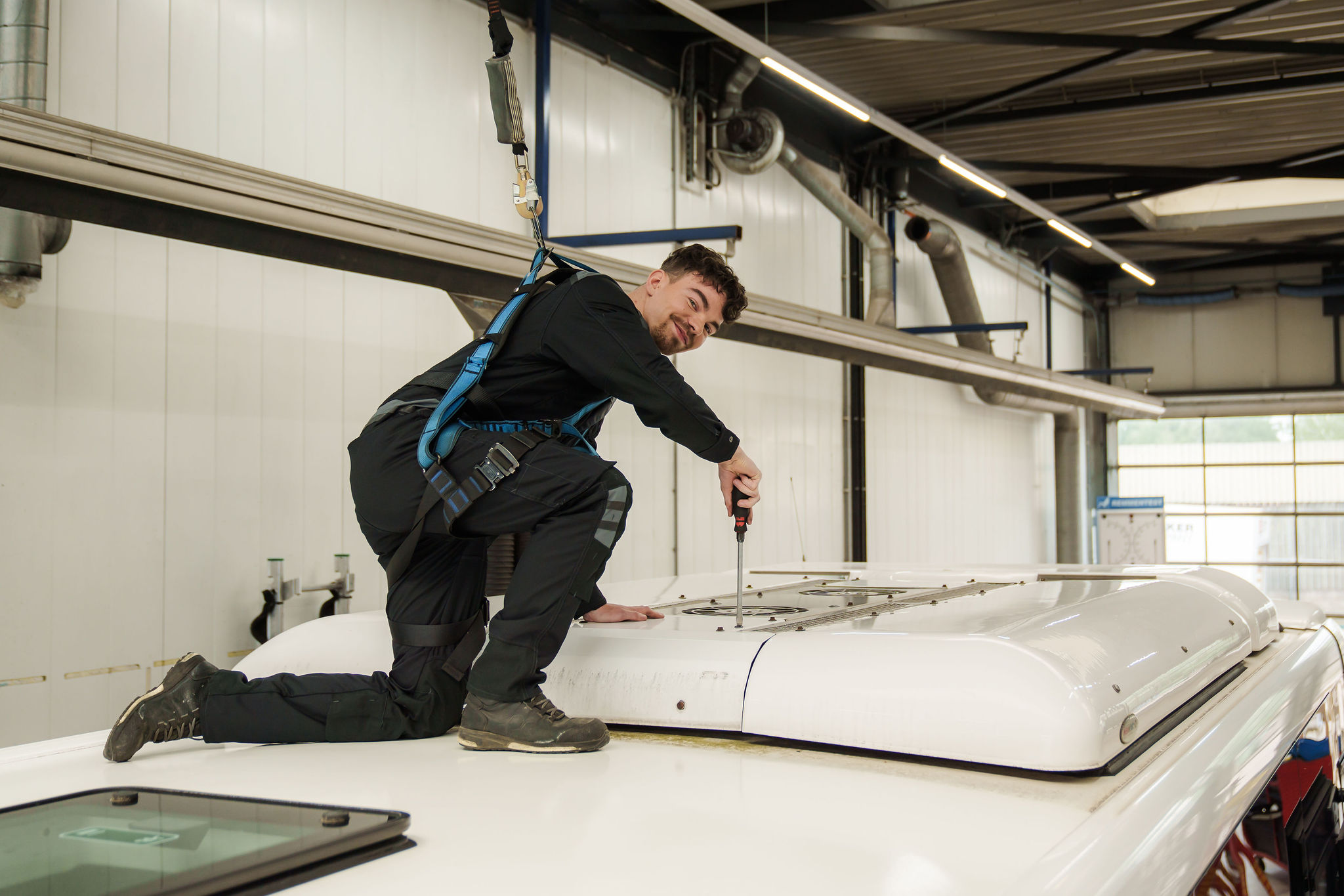 Een monteur van Arriva in veiligheidsharnas werkt op het dak van ene bus in een werkplaats en kijk lachend in de camera.