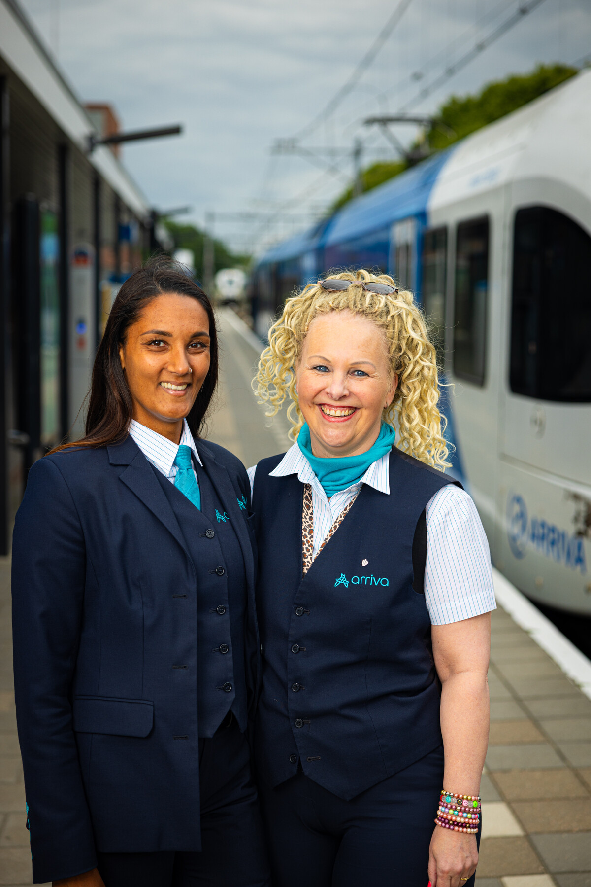 Twee vrouwelijke Stewards BOA-OV van Arriva in uniform staan glimlachend naast elkaar op een perron, met een trein op de achtergrond.