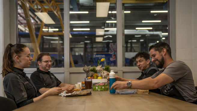 Vier monteurs, drie mannen en een vrouw, zitten aan tafel in de pauzeruimte van de werkplaats en eten en drinken samen