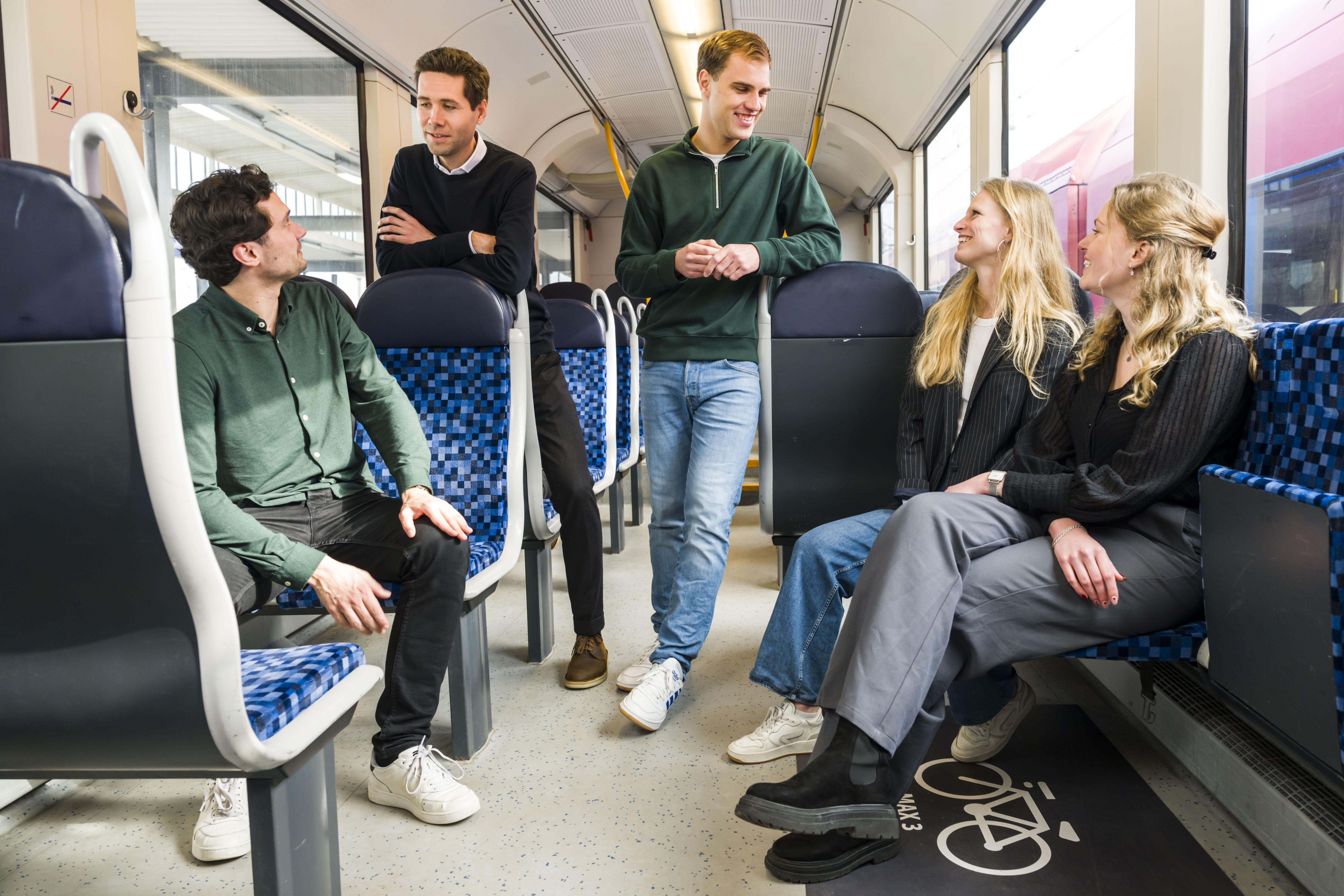 Vijf jonge trainees staan en zitten in treinwagon, levendig discussiërend, de binnenkant van de moderne Arriva-trein zichtbaar.