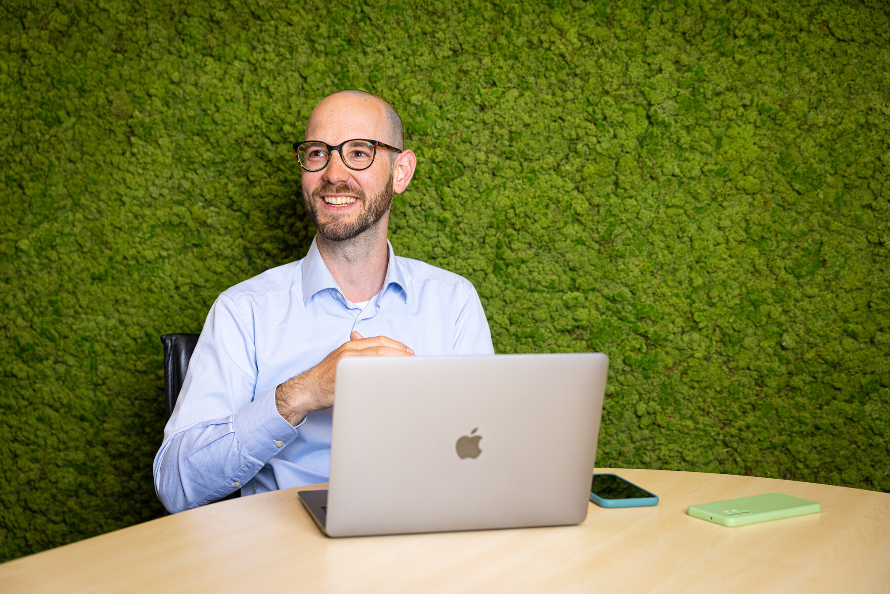 Een jonge mannelijke projectmanager met een bril zit aan een tafel met zijn laptop voor een groene achtergrond.