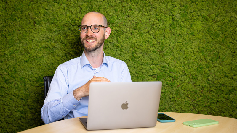 Een jonge mannelijke projectmanager met een bril zit aan een tafel met zijn laptop voor een groene achtergrond.