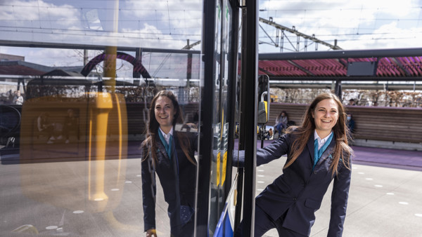 Jonge vrouwelijke buschauffeur draagt een Arriva-uniform en hangt uit de ingang van haar bus.