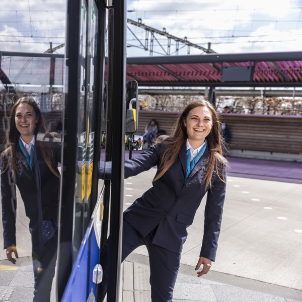Jonge vrouwelijke buschauffeur draagt een Arriva-uniform en hangt uit de ingang van haar bus.