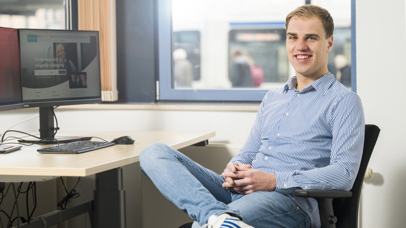 Jonge man in casual kleding zit glimlachend in bureaustoel voor een bureau met computer, achtergrond toont raamuitzicht op trein.