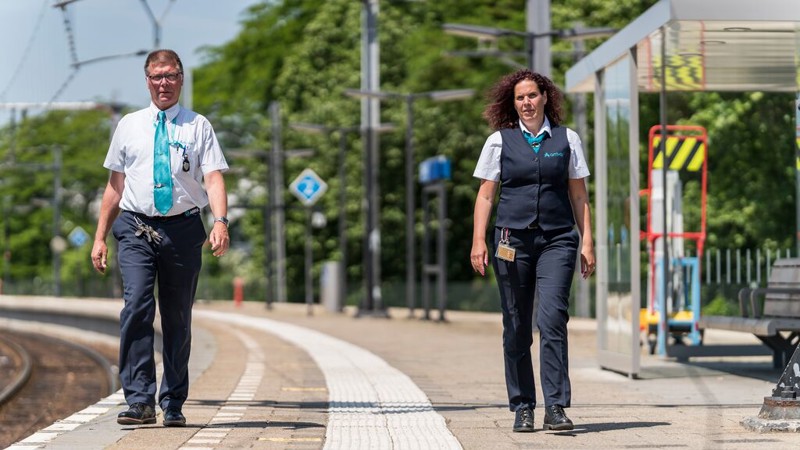 Een mannelijk en vrouwelijke steward van Arriva, lopen over een zonnig treinperron met bomen op de achtergrond.