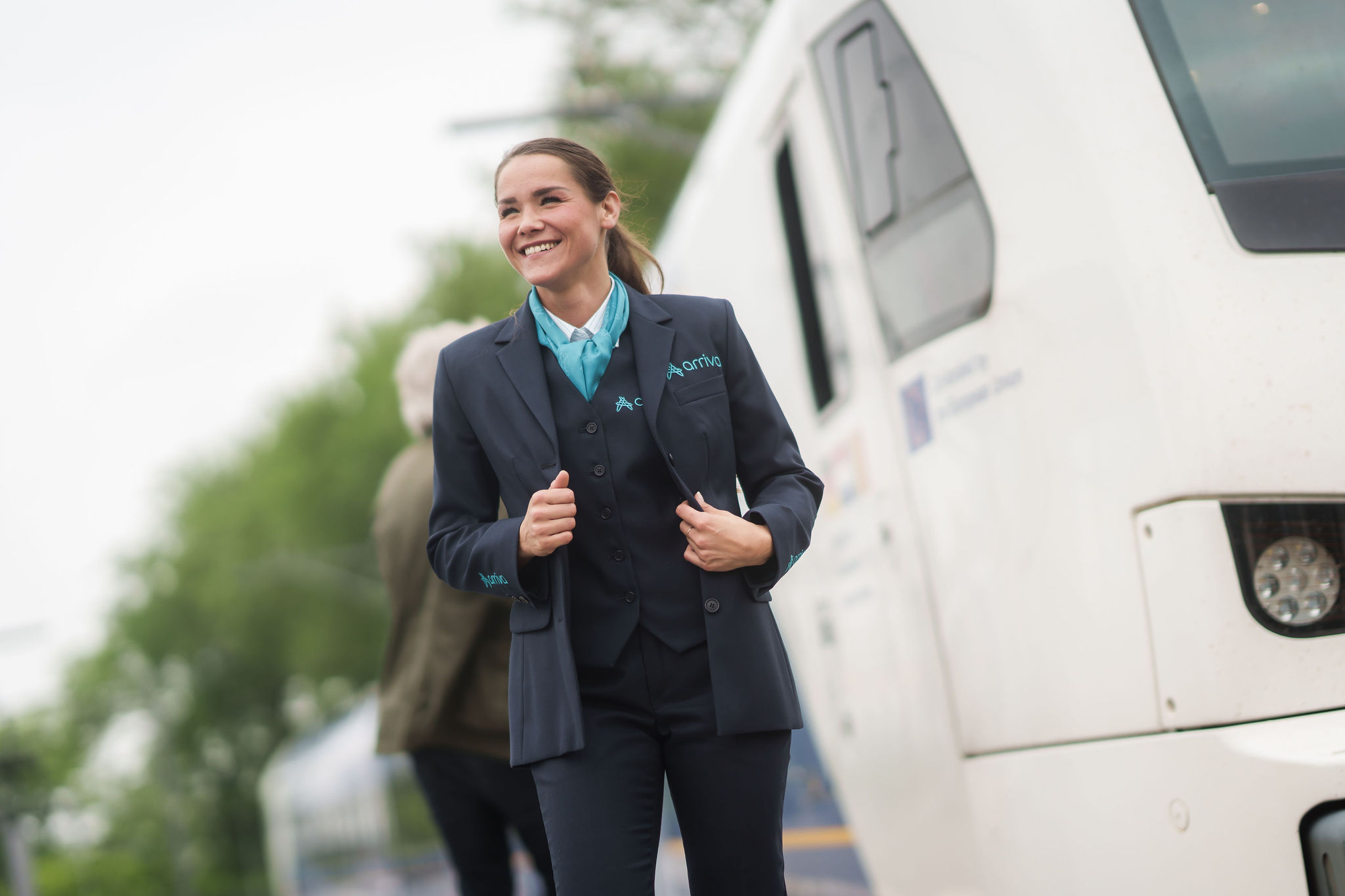 Machinist van Arriva lacht en trekt haar uniform recht, achter haar zie je een reiziger en een trein.