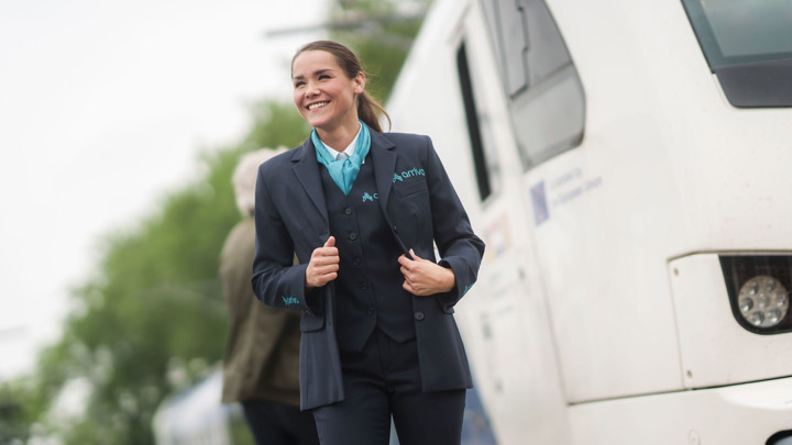 Machinist van Arriva lacht en trekt haar uniform recht, achter haar zie je een reiziger en een trein. Machinist van Arriva lacht en trekt haar uniform recht, achter haar zie je een reiziger en een trein.
