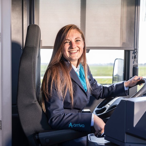 Jonge, vrouwelijke buschauffeur in uniform zit lachend achter het stuur van Arriva-bus.