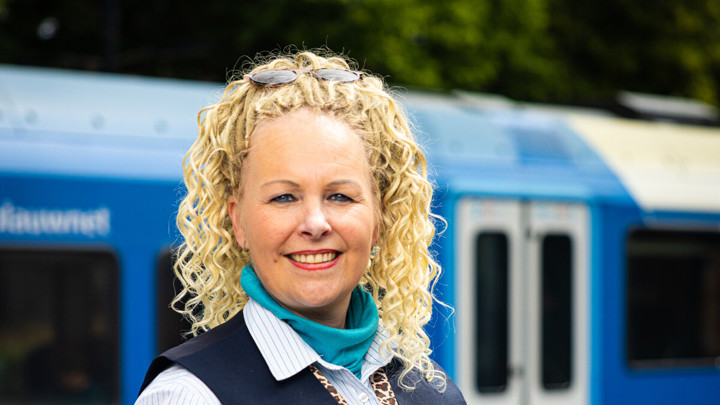 Een Steward BOA-OV staat glimlachend op een perron, met een blauwe trein op de achtergrond. Een Steward BOA-OV staat glimlachend op een perron, met een blauwe trein op de achtergrond.