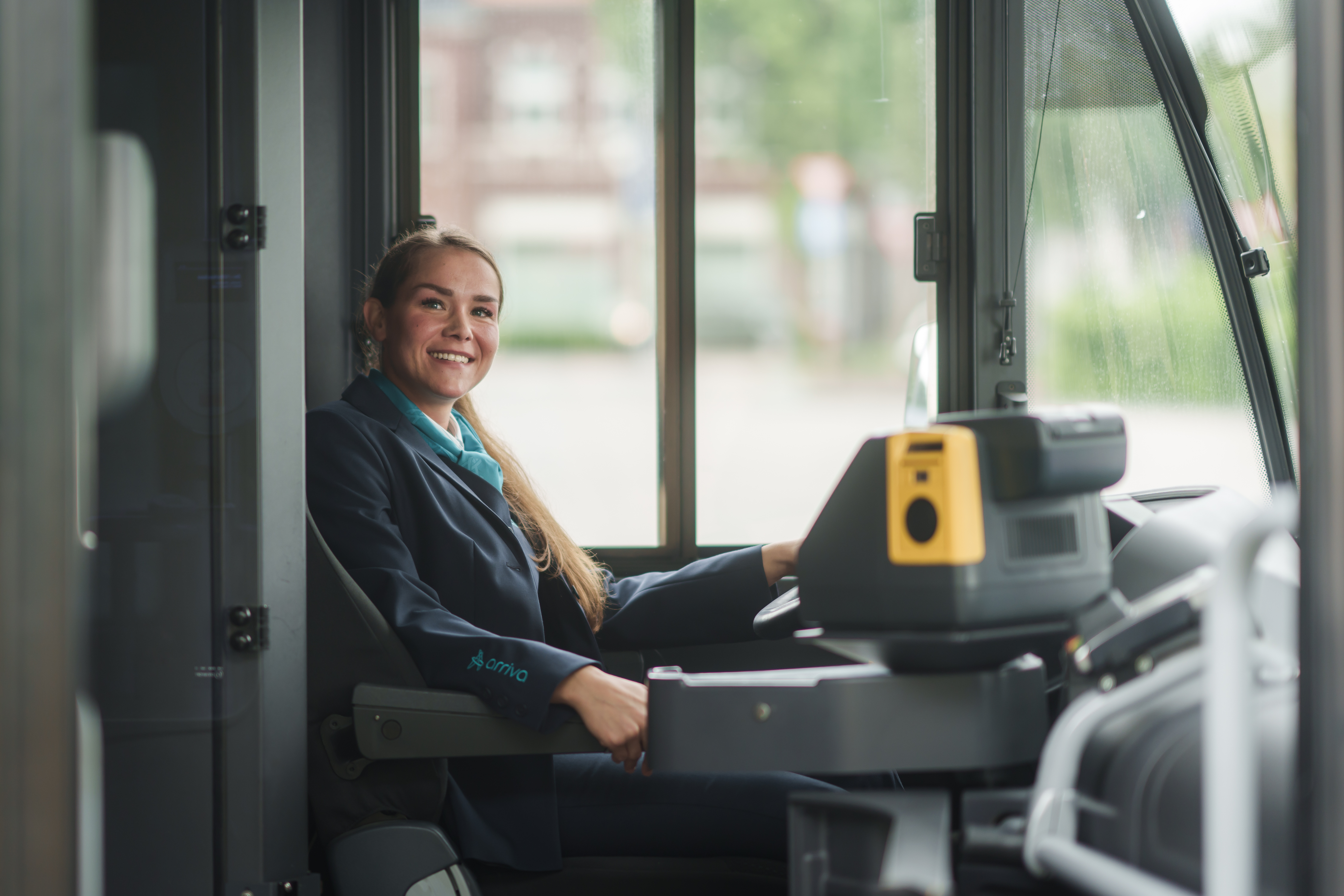 Buschauffeur zit achter stuur van een bus en kijkt lachend opzij in de camera.